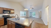 Located at 10550 River Oaks Dr in Perrysburg, Ohio, this well-appointed single-story home offers 2 bedrooms and 2 bathrooms within a thoughtfully designed floor plan. The heart of the home is an open-concept living, dining, and kitchen area anchored by soaring vaulted ceilings and warm hardwood-style flooring. The kitchen features a central island with a double sink and granite-look countertops, warm wood cabinetry, black appliances, and a chandelier overhead, all flowing seamlessly into a bright, carpeted living room with a ceiling fan and sliding glass door access to the outdoors. The primary bedroom is generously sized, complete with a ceiling fan, plush carpet, and access to a private bathroom featuring a walk-in shower with grab bars and a built-in seat. Additional closet space is abundant throughout, including a spacious walk-in closet with wire shelving. The second bedroom offers soft carpet and neutral tones. A dedicated laundry room includes a utility sink, washer/dryer hookups, and overhead shelving. The pantry and hallway provide efficient circulation, and the attached garage adds everyday convenience. The exterior presents tidy landscaping with decorative stone beds and classic vinyl siding.