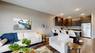 This single-floor unit offers a thoughtfully designed open-concept layout that seamlessly connects the living area and kitchen, creating an inviting and functional living space. The living area features rich, dark hardwood-style flooring, neutral gray walls, and is anchored by a comfortable seating arrangement. A round wooden coffee table, patterned area rug, and nature-inspired wall art contribute to a relaxed, contemporary aesthetic. A floor lamp adds a warm accent to the space.

The kitchen is well-equipped with dark espresso-toned cabinetry extending to the ceiling, granite-style countertops, and a full suite of stainless steel appliances including a range, microwave, and refrigerator. A peninsula with white bar stools serves as both a prep surface and casual dining area, reinforcing the open flow between kitchen and living spaces.

The bedroom is carpeted and bathed in natural light through multiple large windows. Its generous wall space and neutral palette provide a calm, restful environment. The bathroom features dark wood cabinetry, a white vanity with a large mirror, a tub-shower combination with a light curtain, and consistent hardwood-style flooring that ties the unit together cohesively.
