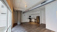 This contemporary loft-style unit showcases an industrial-inspired design that seamlessly blends modern finishes with urban character. The open-plan living area is defined by rich hardwood flooring, soaring ceilings with exposed silver ductwork, and structural cylindrical columns that add architectural interest. Track lighting runs along the ceiling, complementing the bright, airy atmosphere throughout the space.

The kitchen is well-appointed with crisp white shaker cabinetry, granite-look countertops, and a full suite of stainless steel appliances including a refrigerator, range, microwave, and dishwasher. The layout is efficient and functional, offering ample storage and counter space for everyday use.

The living area benefits from expansive floor-to-ceiling windows that frame impressive city skyline views, flooding the interior with natural light and creating a strong connection to the urban environment outside. A glass door provides direct access to an outdoor space, further enhancing the openness of the unit.

The bathroom features a clean, accessible design with a walk-in tiled shower, light marble-style tile flooring, and a drop-ceiling finish. Grab bars and an open layout make the space practical and easy to navigate. Overall, the unit delivers a compelling combination of industrial aesthetics, modern conveniences, and striking cityscape views.