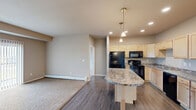 Located at 2829 27th St W in Williston, North Dakota, this 2-bedroom, 2-bathroom single-floor residence offers a practical and well-appointed layout. The open-concept living and dining area flows seamlessly into the kitchen, where light maple cabinetry, granite-style countertops, a central peninsula with pendant lighting, and a full suite of black appliances — including a refrigerator, electric range, microwave, and dishwasher — create a functional and attractive space. Dark hardwood-style flooring anchors the kitchen and dining area, while plush carpet extends throughout the living room and bedrooms. The living room is generously sized, featuring a sliding glass door with vertical blinds that invites natural light and provides outdoor views, along with a wall-mounted AC unit and baseboard heating. The primary bedroom offers comfortable proportions with its own closet and direct access to a bathroom featuring granite-style countertops and light wood cabinetry. The second bedroom is bright and carpeted with a large window. Both bathrooms are well-equipped, with one featuring a full tub-shower combo. Additional closets and a pantry provide ample storage throughout. Neutral warm-toned walls and consistent finishes give the space a cohesive, move-in-ready feel.