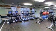 This fitness center is a dedicated single-floor space designed to support a well-rounded exercise routine. The room is outfitted with a row of modern treadmills positioned along the windowed wall, each equipped with individual display consoles for tracking workout metrics. Large windows fitted with horizontal blinds allow natural light to fill the space while maintaining privacy, creating a bright and energizing atmosphere.

The flooring consists of durable, speckled rubberized material — a practical and safety-conscious choice suited for high-traffic fitness environments. A full-length mirror spans a portion of one wall, visually expanding the room and allowing users to monitor their form during workouts.

Additional equipment includes exercise balls in multiple sizes and a weight bench, offering variety for strength and flexibility training. Overhead fluorescent lighting supplements the natural light, ensuring the room remains well-lit at all hours. The walls feature a distinctive striped accent design in navy and tan tones, adding a sporty, dynamic character to the interior. Ventilation is provided through ceiling-mounted vents, keeping the environment comfortable during use. The overall layout is open and functional, offering ample space to move freely between equipment.