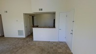 This single-floor space is designed with an open-concept layout that promotes a sense of spaciousness and functionality. The main living area features a combination of tile flooring near the entryway and carpeting extending through the remainder of the room, creating distinct zones within the open floor plan. Soaring ceilings add a dramatic vertical dimension to the space, enhancing the overall sense of airiness.

A defining architectural feature is the pass-through kitchen bar, finished with a stone-look countertop and supported by a clean white base. This peninsula-style counter connects the kitchen to the living area, making it ideal for casual dining or entertaining. The kitchen, visible through the pass-through opening, accommodates a refrigerator and provides additional workspace and storage.

The living area includes multiple doorways leading to additional rooms, offering flexible access throughout the unit. White interior doors and neutral wall tones maintain a clean, cohesive aesthetic. HVAC vents are positioned both near the floor and higher on the walls, ensuring efficient climate control. The overall palette is light and neutral, providing a versatile backdrop for various furnishing styles.