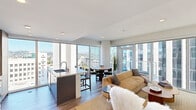 This well-appointed high-rise unit offers a thoughtfully designed open floor plan spanning a generous single level, with two bedrooms and two bathrooms. Floor-to-ceiling windows wrap around the main living areas, flooding the space with natural light and framing panoramic views of the surrounding cityscape and distant mountains.

The open-plan living, dining, and kitchen area serves as the heart of the unit. The kitchen features flat-front cabinetry in a warm wood-tone finish, stainless steel appliances including a gas range and French-door refrigerator, and a sleek island with bar seating. The dining area sits adjacent, benefiting from the same expansive window views, while the living room offers direct access to the exterior through glass doors.

The primary bedroom is a well-proportioned retreat with large windows overlooking the city skyline. The second bedroom, equally bright, functions flexibly as a sitting room or guest space. Both bathrooms are cleanly finished, with one featuring a soaking tub and marble-style tile surround. Light wood-tone flooring runs throughout, complementing the unit's contemporary, minimalist aesthetic and reinforcing a cohesive, modern ambiance.