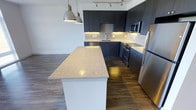 This single-floor space showcases a thoughtfully designed kitchen that serves as a central feature of the layout. The kitchen is anchored by a generously sized island with a light-toned granite countertop, offering ample workspace and a natural gathering point. Dark, flat-front cabinetry extends from floor to ceiling, providing substantial storage while lending a sleek, contemporary aesthetic. Under-cabinet lighting illuminates the countertops below, enhancing both functionality and visual warmth.

The cabinetry is paired with a classic white tile backsplash that creates a clean contrast against the darker tones. A full suite of stainless steel appliances — including a refrigerator, stove, built-in microwave, and dishwasher — reinforces the kitchen's modern sensibility. A stainless steel sink is integrated seamlessly into the perimeter countertop.

Overhead, industrial-style pendant lights hang above the island, complemented by a track lighting system mounted to the ceiling. The rich, dark hardwood flooring runs throughout, grounding the space with warmth and continuity. The overall ambiance is clean, modern, and functional, with a cohesive material palette throughout.