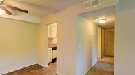 This single-floor residential unit offers a practical and straightforward layout designed for comfortable everyday living. The space is characterized by warm, neutral-toned walls that create a welcoming ambiance throughout. A central hallway connects the various areas of the unit, providing a clear sense of flow and easy navigation between rooms.

The kitchen is visible through an open archway and is outfitted with white cabinetry, a dark countertop, and built-in appliances including a dishwasher, making it a functional space for meal preparation. The cabinetry provides ample storage, and the layout appears efficient for everyday use.

The flooring transitions between durable hardwood-style planks in the main living and kitchen areas to soft carpeting along the hallway, adding warmth and texture as you move through the space. The textured ceiling is consistent throughout and features centrally placed light fixtures that provide even, ambient illumination. Overhead HVAC venting ensures climate control is integrated seamlessly into the design.

Overall, the unit presents a clean, functional living environment with a cohesive color palette of warm neutrals, practical finishes, and a straightforward layout that makes efficient use of the available space.