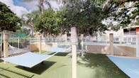 This single-level outdoor recreational space offers an engaging environment designed for active use and leisure. The area is enclosed by a white metal perimeter fence, providing a secure and defined boundary while maintaining an open, airy feel. Synthetic turf covers the ground throughout, offering a clean and resilient surface suited for a variety of activities.

A standout feature of the space is its rope climbing structure — sturdy vertical posts wrapped with thick braided rope — which serves as an interactive element for physical engagement. Alongside this, a slanted ramp or climbing board adds further variety to the recreational offerings. The layout is thoughtfully designed to encourage movement and play.

Mature trees, including broad-canopied shade trees and tall palms, are planted within and around the space, lending natural beauty and generous shade on sunny days. The lush canopy overhead creates a pleasant, park-like atmosphere that softens the built environment. Lamp posts are also present, suggesting the area is accessible and usable beyond daylight hours.

The surrounding architecture, partially visible beyond the fence, reflects a warm residential character. Overall, this outdoor space strikes a balance between structured activity zones and natural, green surroundings, making it a welcoming and functional recreational destination.