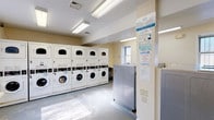The Princeton Gardens laundry facility is a well-organized, single-floor communal space designed to serve residents with efficiency and ease. The room is outfitted with an extensive lineup of stacked washer and dryer units, numbered for easy identification, arranged along one wall to maximize available floor space. The machines are modern, front-loading models that accommodate a range of laundry needs.

The interior features neutral, light-toned walls paired with a durable, easy-to-clean floor, creating a clean and functional environment. Overhead fluorescent lighting ensures the space is well-lit throughout. Multiple windows allow for natural light to supplement the artificial lighting, contributing to a bright and airy atmosphere.

A wall-mounted folding table provides a convenient surface for sorting and folding laundry, while a cork bulletin board serves as an information hub for residents. Informational signage is posted throughout the room, including usage instructions and community guidelines. A stainless steel utility cabinet offers additional storage or workspace.

The layout provides generous open floor space, allowing multiple users to move comfortably within the room simultaneously. The facility also includes a separate entry door and clearly marked exit, ensuring safe and straightforward access. Overall, this laundry room is a practical and well-maintained amenity for Princeton Gardens residents.