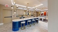 Located at 805 N Olive Ave in West Palm Beach's vibrant Nora District, Tortoise One is a thoughtfully designed amenity lounge spanning a generous single floor. The entry hallway welcomes guests with a patterned tile floor, a built-in upholstered bench, lush greenery, and a textured metallic ceiling that sets an immediately sophisticated tone. Floor-to-ceiling glass doors flood the entry with natural light and offer views of surrounding palm trees. The kitchen is anchored by a striking cobalt blue island with a thick marble-topped counter and gold-accented barstools, complemented by warm cabinetry, a mosaic tile backsplash, and a sculptural multi-globe pendant lighting arrangement. An adjacent elongated bar counter with matching stone surface provides additional seating. The living room is a vibrant artistic focal point, featuring a mirrored ceiling that reflects a colorful mural, a plush sectional sofa, and a curated gallery wall of bold contemporary paintings framed by large windows overlooking the outdoors. The intimate dining nook offers banquette seating in rich blue fabric beneath a vivid red statement artwork. The game room rounds out the space with a full-size billiard table, cove-lit tray ceiling, a wall-mounted television, and additional lounge seating — creating a dynamic and inviting social environment throughout.