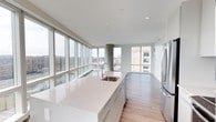 This contemporary 3-bedroom, 3-bathroom residence offers a seamless blend of modern design and urban living. The open-concept layout is anchored by a sleek, modern kitchen featuring an expansive quartz island with an integrated sink, flat-front cabinetry, and stainless steel appliances. Light hardwood flooring flows throughout the space, complementing the clean, neutral color palette. One of the most striking features of this residence is the dramatic floor-to-ceiling window system that spans the main living area, flooding the interior with abundant natural light and framing panoramic views of the surrounding cityscape. A structural concrete column adds an industrial accent that contrasts elegantly with the refined finishes throughout. The open living and dining areas benefit directly from the wall of glass, creating an airy, elevated atmosphere with a strong connection to the outdoors. The three bedrooms and three full bathrooms provide generous accommodation, making this an ideal space for comfortable everyday living. The overall ambiance is bright, sophisticated, and modern, with thoughtful architectural details that enhance both function and aesthetic appeal throughout.