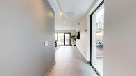 Located at 1275 N Santa Fe Dr in Denver's vibrant arts district, this contemporary communal lounge offers a well-appointed, open-plan layout across a single floor. The space is defined by large-format terrazzo-style tile flooring that extends throughout, contributing to a clean, polished aesthetic. The main living area is anchored by a set of rich brown leather sofas and armchairs arranged around a sculptural glass-top coffee table, all set atop a textured area rug. Recessed ceiling lighting and structured ceiling coffers add architectural interest while keeping the atmosphere bright and welcoming. Large windows and a full-height glass sliding door flood the interior with natural light and open directly onto an outdoor patio area, offering views of a pool and surrounding rooftop space. A wet bar with stone tile backsplash and gray cabinetry lines one wall, accompanied by a coffee station, making the space well-suited for social gatherings. A round dining table with sleek chairs provides a casual meeting or dining area. Curated wall art and a wall-mounted flat-screen television add personality, while a hallway with elevator access ensures seamless connectivity. The overall ambiance is modern, social, and inviting.