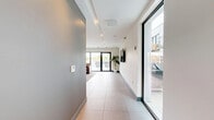 Located at 1275 N Santa Fe Dr in Denver's vibrant arts district, this contemporary communal lounge offers a well-appointed, open-plan layout across a single floor. The space is defined by large-format terrazzo-style tile flooring that extends throughout, contributing to a clean, polished aesthetic. The main living area is anchored by a set of rich brown leather sofas and armchairs arranged around a sculptural glass-top coffee table, all set atop a textured area rug. Recessed ceiling lighting and structured ceiling coffers add architectural interest while keeping the atmosphere bright and welcoming. Large windows and a full-height glass sliding door flood the interior with natural light and open directly onto an outdoor patio area, offering views of a pool and surrounding rooftop space. A wet bar with stone tile backsplash and gray cabinetry lines one wall, accompanied by a coffee station, making the space well-suited for social gatherings. A round dining table with sleek chairs provides a casual meeting or dining area. Curated wall art and a wall-mounted flat-screen television add personality, while a hallway with elevator access ensures seamless connectivity. The overall ambiance is modern, social, and inviting.