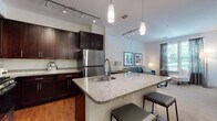 This single-floor residence offers a thoughtfully designed open-concept layout that seamlessly connects the kitchen, dining, and living areas. The kitchen is a standout feature, showcasing rich dark-stained cabinetry that extends to the ceiling, granite countertops, and a full suite of stainless steel appliances including a gas range, built-in microwave, and refrigerator. A large central island with an undermount sink and bar seating serves as both a functional workspace and a social hub. Pendant lighting and track lighting illuminate the kitchen and flow naturally into the open living space. The living room is bright and welcoming, benefiting from multiple windows that allow abundant natural light and offer outdoor views. Warm hardwood flooring anchors the kitchen and entry areas, while plush carpet extends through the living room and bedroom. The bedroom is a serene retreat, featuring large windows with outdoor views, ample space for a full bedroom suite, and a calm, neutral color palette. The bathroom is clean and functional, finished with large-format tile flooring, a dark-stained vanity with a light countertop, and a full tub-shower combination. Throughout the residence, a cohesive design language of neutral tones and contemporary finishes creates a polished, comfortable living environment.