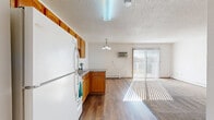 This studio in Fargo, North Dakota offers a well-organized, single-floor layout that maximizes every square foot. The combined kitchen and living area serves as the heart of the space, featuring warm honey-toned wood cabinetry, laminate countertops, a double-basin sink, and white appliances including a refrigerator, range, and microwave. Hardwood-style flooring runs throughout the kitchen area, lending a clean and cohesive look, while a pendant light fixture adds a warm accent overhead. A textured ceiling and bright white walls keep the space feeling open and airy. The living area transitions seamlessly from the kitchen, offering carpeted flooring and abundant natural light streaming through a large sliding glass door fitted with vertical blinds. The door opens onto a private balcony, providing pleasant outdoor views of the surrounding grounds. Baseboard heating runs along the walls throughout the unit, ensuring comfortable warmth year-round, complemented by a wall-mounted air conditioning unit for warmer months. The bathroom is clean and functional, featuring a full tub-shower combination with white tile surround. A dedicated closet provides practical storage. This studio offers a straightforward, comfortable layout well-suited for efficient everyday living.