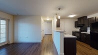 This single-floor residence offers a thoughtfully designed open-concept layout that seamlessly connects the living and kitchen areas. Rich, dark hardwood-style flooring runs throughout the main living spaces, creating a warm and cohesive aesthetic. The kitchen is well-equipped with dark espresso cabinetry, a suite of black appliances including a refrigerator, range, and dishwasher, and a light-toned countertop that extends into a peninsula breakfast bar. Pendant lighting above the peninsula adds a refined touch, while ample cabinet storage lines both sides of the galley-style kitchen. A hallway leads to the private areas of the residence, featuring multiple paneled doors and convenient closet access. The bedroom is carpeted for comfort and receives natural light through a generously sized window with blinds. Two bathrooms serve the space: one features a full tub-shower combination with chrome fixtures and clean white surroundings, while the second includes a vanity with a dark cabinet base, a mirror, and access to an adjacent shower area. Throughout the residence, white walls and neutral tones create a bright, clean atmosphere that lends itself well to a variety of interior styles.