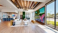 Located at 2500 Conserve Circle in Apopka, Florida, this single-story clubhouse for Novo Avian Pointe offers an expansive, open-concept layout spanning nearly 3,000 square feet of thoughtfully designed community space. The interior is defined by striking rustic wood beam ceilings, light hardwood flooring, and floor-to-ceiling black-framed glass windows that flood the space with natural light and provide views of the surrounding community.

A dramatic focal point of the main living area is an oversized nature-inspired mural depicting Florida wildlife, including herons and roseate spoonbills, framed within a reclaimed wood surround. A sculptural multi-arm chandelier anchors the lounge area, which is appointed with contemporary furnishings, a cozy seating arrangement, and layered rugs. A dedicated workspace with a desk and green velvet seating offers a functional retreat within the open floor plan.

The clubhouse also features a fully equipped kitchen with soft gray cabinetry, quartz countertops, a large island with bar seating, stainless steel appliances, and a wood plank ceiling with Edison-style pendant lighting. An adjacent dining area with a long communal table and a lounge zone with tray ceilings and ambient lighting complete the space. A welcoming entry area with a sofa and digital display screen rounds out this well-appointed community hub.