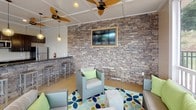 This single-floor amenity space is thoughtfully designed to serve as a multifunctional gathering and entertainment hub. The interior is organized into distinct functional zones that flow seamlessly together, creating an inviting atmosphere for group activities and socializing.

The lounge area is anchored by a striking stacked-stone accent wall, which provides a bold textural contrast to the otherwise light, neutral walls and coffered ceiling. A wall-mounted flat-screen TV is framed within the stonework, making it a natural focal point. Wicker seating with green accent cushions is arranged around a colorful blue and green patterned area rug, enhancing the relaxed, coastal ambiance. Ceiling fans throughout contribute to comfortable airflow.

Large floor-to-ceiling windows and glass panels flood the space with natural light and offer unobstructed views of the surrounding outdoor area, which appears to include a pool and landscaped grounds. A separate dining zone accommodates a long rectangular table with bench seating, ideal for group meals.

The kitchenette is well-appointed with dark espresso cabinetry, dark granite countertops, stainless steel appliances including a refrigerator and microwave, and a bar-height counter with stools — providing ample functionality for catered events or casual use.