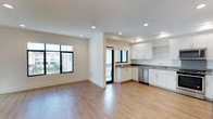 This single-floor space features an open-concept layout that seamlessly connects the living area and kitchen, creating a sense of openness and ease of movement throughout. The living area is bathed in natural light, courtesy of large black-framed windows that frame views of the surrounding neighborhood and greenery outside. Recessed lighting in the white ceiling supplements the natural light, ensuring the space remains well-lit throughout the day.

The kitchen is thoughtfully designed with white shaker-style cabinetry, offering ample storage through both upper and lower cabinets, complemented by open shelving above. Dark countertops provide a stylish contrast to the white cabinetry, while a stainless steel dishwasher and sink add modern, functional elements. A second window positioned in the kitchen further enhances the brightness of the cooking area.

Consistent light-toned hardwood-style flooring runs throughout the entire space, adding warmth and cohesion to the open layout. Clean white walls and minimal detailing contribute to a fresh, contemporary aesthetic, making this a versatile and welcoming living environment.