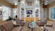 This single-story amenity facility is thoughtfully designed to serve as a central gathering hub. The main lounge area makes an immediate impression with its coffered ceiling detail, a striking multi-pendant chandelier, and a ceiling fan overhead. Built-in shelving units with recessed lighting flank a wall-mounted television, providing both display space and a focal point for the seating area. Patterned accent chairs surround a glass-top coffee table atop a plush area rug, while large windows allow natural light to fill the room and offer views of the surrounding greenery.

Adjacent to the lounge is a dining area with a round glass table, positioned near a compact kitchenette. The kitchen features warm honey-toned cabinetry, stone-look countertops, a double sink, stainless steel appliances, and generous storage — making it well-suited for hosting small gatherings or events.

The fitness center occupies a dedicated section of the facility and is outfitted with rubber flooring, a multi-station cable machine, elliptical trainers, stationary bikes, and additional cardio equipment. Crown molding, ceiling fans, and large windows with natural light give the workout space a bright, polished feel. A separate door provides direct outdoor access, enhancing ventilation and convenience.