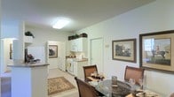 This single-floor residence offers an open and functional layout where the kitchen and dining area flow seamlessly together. The kitchen is outfitted with white cabinetry, white appliances including a gas range, built-in microwave, and a full-size refrigerator. Decorative baskets atop the upper cabinets add a homey touch, while the countertops provide ample prep space. A flush-mount ceiling light brightens the area effectively. The dining space accommodates a glass-top table surrounded by upholstered chairs, making it well-suited for everyday meals or casual entertaining. Framed artwork on the neutral-toned walls adds warmth to the space. Light-colored flooring runs throughout, complemented by an area rug in the dining zone that helps define the space. A doorway visible in the background suggests additional rooms beyond, indicating a connected and practical floor plan. The overall aesthetic is clean, neutral, and move-in ready, with a simple, understated design that prioritizes functionality and livability.