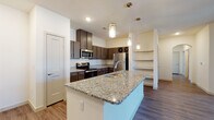 This single-floor residence offers a thoughtfully designed open-concept layout that seamlessly connects the kitchen and living areas. The kitchen is a standout feature, showcasing granite countertops, dark espresso-toned shaker cabinetry, and a full suite of stainless steel appliances including a refrigerator, electric range, and microwave. A large kitchen island with an undermount sink and elegant pendant lighting serves as both a functional workspace and a natural gathering point. A built-in wine rack and open shelving unit add character and additional storage. The living area is spacious and bright, featuring wood-look flooring and a ceiling fan, creating a comfortable and open atmosphere. The bedrooms are carpeted for warmth and comfort, each equipped with ceiling fans and windows that allow natural light and exterior views. The primary bathroom includes a dual-sink granite vanity, wood-look flooring, and a walk-in glass-enclosed shower. A secondary full bathroom features a tub-shower combination with tile surround, a granite vanity, and matching dark cabinetry. Throughout the space, consistent finishes including granite surfaces, dark cabinetry hardware, and wood-look flooring create a cohesive and modern aesthetic.