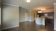 This single-floor residence offers an open-concept layout that seamlessly connects the living area and kitchen. Dark hardwood-style flooring extends throughout, providing a rich contrast against the light gray and neutral-toned walls accented with white crown molding and baseboards. The living area is generously proportioned, offering flexible space for various furniture arrangements, and is lit by a flush-mount ceiling light fixture. The front entry features a solid white door, contributing to a clean, welcoming aesthetic.

The kitchen is positioned to the right and distinguished by dark espresso cabinetry, a stainless steel refrigerator, and a curved breakfast bar finished with granite-style countertops. A pendant light fixture hangs above the bar, adding a touch of warmth to the space. A doorway visible beyond the kitchen suggests access to additional rooms or utility spaces. The overall ambiance is modern and practical, with a neutral color palette and consistent finishes that create a cohesive, move-in-ready environment throughout the unit.