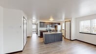 This single-floor residence offers a clean, modern layout designed for comfortable everyday living. The heart of the home is an open-concept kitchen, anchored by a center island with an undermount sink and ample counter space finished in neutral tones. Dark espresso cabinetry lines the walls, providing generous storage, while stainless steel appliances — including a range, built-in microwave, refrigerator, and dishwasher — lend a polished, contemporary feel. Light hardwood-style flooring flows seamlessly throughout the space, unifying each area with a warm, cohesive aesthetic.

Adjacent to the kitchen, a hallway connects to additional rooms, including a laundry area visible in the background. The living area is generously sized and filled with natural light from a sliding glass door that opens onto a private balcony overlooking mature trees. A wall-mounted mini-split unit ensures year-round climate comfort in this open space.

The bathroom features a dual-vanity setup with dark cabinetry, neutral countertops, and a large mirror, complemented by a separate water closet for added privacy. Windows throughout the unit allow natural light to brighten each room. The overall ambiance is fresh, neutral, and move-in ready, with a practical yet attractive design suited to modern living.