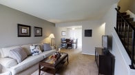 This two-floor residence offers a thoughtfully arranged living space characterized by a seamless flow between its main gathering areas. The living room serves as a comfortable focal point, furnished with a neutral-toned sofa accented by decorative pillows and anchored by a dark wood coffee table with glass insets. Warm lighting from a classic floor lamp and wall-mounted sconce creates an inviting atmosphere, while framed artwork adds a touch of refined character to the softly painted walls.

The living area opens directly into a dedicated dining space, distinguished by a chandelier overhead and a full dining set suitable for seated gatherings. Vertical blinds on the adjacent window allow for adjustable natural light, contributing to the room's bright and airy feel. Plush wall-to-wall carpeting runs throughout both spaces, adding warmth and continuity to the overall design.

The neutral color palette of whites, grays, and beiges gives the interior a clean, cohesive aesthetic. A media console near the living area provides practical storage and display space. Spanning two floors, this residence offers additional living space beyond what is visible, making it well-suited for a variety of lifestyle needs.