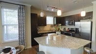 This single-floor space showcases a well-appointed kitchen designed with both functionality and aesthetics in mind. The kitchen is anchored by a large curved island topped with light-toned granite countertops that provide generous workspace and a striking visual contrast against the rich, dark espresso cabinetry. The cabinetry extends from floor to ceiling, offering abundant storage throughout the space.

Stainless steel appliances, including a range with electric cooktop and an over-the-range microwave, lend a sleek, contemporary feel. A stainless steel sink is positioned beneath a window, allowing natural light to filter in and brighten the workspace. The window is dressed with a patterned Roman shade that complements the overall design palette.

Overhead, a combination of track lighting and two globe-style pendant lights hung above the island provides layered, practical illumination. The flooring is a warm hardwood tone that grounds the space and coordinates with the bold cabinetry. Patterned drapery along one wall adds texture and depth, contributing to a cohesive and polished interior aesthetic throughout the kitchen area.