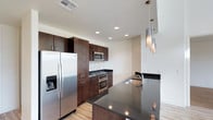 This contemporary two-floor space is designed with modern finishes and thoughtful detail throughout. The kitchen serves as a standout feature, showcasing rich dark-toned cabinetry that extends to the ceiling, providing generous storage. A full suite of stainless steel appliances — including a French door refrigerator with an ice and water dispenser, a built-in microwave, a gas cooktop, and a wall oven — delivers both functionality and a polished aesthetic. Dark granite countertops provide ample workspace and are complemented by a mosaic tile backsplash that adds subtle texture and visual interest. A central kitchen island, finished with the same granite surface, features an undermount sink with a modern arc faucet, and serves as an ideal space for meal preparation or casual dining. Recessed ceiling lighting throughout the kitchen ensures even illumination, while a cascading set of sleek pendant lights above the island adds a refined, decorative touch. Light wood-toned flooring runs underfoot, creating a warm contrast against the darker cabinetry. The overall ambiance of the space is clean, contemporary, and functional, reflecting a design sensibility that balances practicality with modern style across both floors.