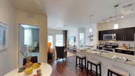 This single-floor residence features an open-concept layout that integrates the kitchen, dining, and living areas into one cohesive and functional space. The kitchen is outfitted with dark cabinetry, granite countertops, stainless steel appliances including a microwave and range, and a double-basin sink. A peninsula with granite overhang accommodates two cushioned bar stools, creating a casual dining option that connects directly to the main living space. A pendant light fixture above the peninsula adds a refined touch.

The living area is generously proportioned, highlighted by rich hardwood-style flooring that extends throughout. Large windows and glass doors at the far end of the living room invite abundant natural light, enhancing the open and airy atmosphere. Track lighting and recessed fixtures illuminate the space effectively.

A separate room, visible through a doorway on the left, appears to serve as a bedroom or sitting area, also benefiting from natural light. Neutral wall tones throughout provide a versatile backdrop, while the smooth white ceilings contribute to the sense of spaciousness. The overall ambiance is modern, bright, and well-appointed.