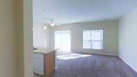 This two-floor space features an open-plan main level where the kitchen and living area flow together seamlessly. The kitchen includes a peninsula counter with warm wood-toned cabinetry, a dishwasher, and a pendant light overhead, providing a functional and connected cooking space. The transition into the living area is smooth, with neutral carpet flooring underfoot and light-toned walls throughout. A ceiling fan with an attached light fixture serves the living area, enhancing airflow and comfort. Two windows, including a large sliding glass door fitted with vertical blinds and a secondary side window, allow generous natural light to pour into the space, creating a bright and airy atmosphere. The textured ceiling adds subtle visual depth. The open layout and abundance of natural light make the main level feel spacious and welcoming, while the neutral color palette throughout provides a versatile backdrop. The property extends across two floors, offering additional living space beyond the main level shown.