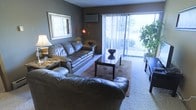 This single-floor space offers a thoughtfully arranged living area that balances comfort and functionality. The living room serves as the centerpiece, featuring plush wall-to-wall carpeting that adds warmth underfoot. Leather seating anchors the space, arranged around a dark coffee table atop a decorative area rug, creating an inviting conversation area. Natural light pours in through a large sliding glass door fitted with vertical blinds, which opens to an outdoor balcony and brings in pleasant views of surrounding greenery. A wall-mounted air conditioning unit ensures year-round comfort. The room is complemented by neutral, dark-toned walls that lend a modern and cozy ambiance. A large framed mirror on the wall enhances the sense of space and adds a decorative touch. Accent lighting from floor and table lamps creates a warm atmosphere during evening hours. A lush indoor plant near the balcony door adds a natural, refreshing element to the interior. The overall design reflects a practical yet stylish layout, well-suited for relaxed everyday living.