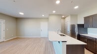 This single-floor unit features a contemporary design highlighted by light-toned luxury vinyl plank flooring, neutral warm-gray walls, and clean white trim throughout. The open-concept layout connects the kitchen and living area seamlessly, creating a bright, cohesive living environment illuminated by recessed lighting.

The kitchen is a standout feature, boasting rich dark-brown shaker-style cabinetry with brushed nickel hardware, crisp white countertops, a peninsula with an undermount sink, and a full suite of stainless steel appliances including a refrigerator, range, and microwave. Ample upper and lower cabinetry provides generous storage.

The living area is spacious and open, flowing toward a private balcony accessible through a glass-panel door flanked by windows dressed with white blinds, bringing in natural light and outdoor views of the surrounding neighborhood. A wall-mounted PTAC unit provides climate control.

The bedroom is comfortably sized with a window framing exterior views and connects to a bathroom featuring a dark-cabinetry vanity, white countertop, large frameless mirror, and bright vanity lighting. An in-unit washer is conveniently positioned nearby, adding everyday practicality to this well-designed space.