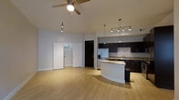 This single-floor space in Dallas, Texas, offers a well-designed open-concept layout that seamlessly blends the living and kitchen areas. The light hardwood flooring extends throughout, creating a warm and cohesive aesthetic. A ceiling fan with integrated lighting anchors the living area, while track lighting above adds additional illumination to the space.

The kitchen is a standout feature, equipped with dark espresso cabinetry that provides generous storage, granite countertops, and a stainless steel refrigerator. A curved kitchen island with a matching granite surface serves as both a functional workspace and a natural divider between the kitchen and living area. Pendant lighting above the island adds a modern, refined touch. The kitchen also features a sink with a window view and additional overhead track lighting to brighten the workspace.

Adjacent to the main living area, sliding panel doors reveal a spacious storage or utility closet, offering practical organization solutions. A dark-finished entry door grounds the space with a bold design element. The overall ambiance is clean, modern, and functional, making this Dallas space an inviting and versatile environment suited for a variety of uses.