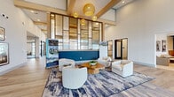 This expansive single-floor amenity and leasing center spans a generously sized footprint, thoughtfully designed to accommodate a variety of functions within a cohesive, modern aesthetic. The space is anchored by a dramatic main lobby featuring soaring double-height ceilings with exposed wood beam detailing, light hardwood-style flooring, and large black-framed windows that draw in abundant natural light. Woven rattan pendant lights hang overhead, casting warm illumination across a curated seating arrangement of rounded upholstered chairs and a sculptural wood coffee table, all set atop a layered abstract area rug.

A bold navy blue accent wall serves as a striking focal point in the lobby, framed by a floor-to-ceiling glass and metal partition that adds architectural depth. Adjacent areas transition seamlessly into more casual lounges with leather sofas, cane-accented chairs, and a statement globe chandelier. A dedicated conference room features a long communal table, blue rolling chairs, patterned carpet, a display screen, and curated wall art.

The booth seating area offers semi-private nooks with cognac leather banquettes, slatted wood dividers, and arc pendant lighting, ideal for focused work or informal gatherings. Throughout, the design balances refined materials with an inviting, community-forward atmosphere.
