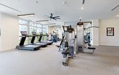 Located at 27077 S Dixie Hwy in Homestead, FL, this single-floor fitness center offers a generously sized, well-appointed exercise facility designed to support a comprehensive range of workout routines. The space is characterized by bright, white walls, light-toned carpet flooring, and modern linear pendant lighting that creates a clean, energizing atmosphere throughout.

The main strength training area is equipped with a robust selection of cable machines, selectorized strength equipment, and a dedicated free weights section featuring a full dumbbell rack lined against large wall mirrors. A power rack with adjustable benches provides space for compound lifts, while motivational wall typography — including bold "DRIVE" and "FOCUS" graphics — reinforces an active environment.

A separate cardio zone lines the perimeter, where floor-to-ceiling windows flood the space with natural light and offer open views of the surrounding landscape. Treadmills with integrated screens and additional cardio machines are positioned to take full advantage of these views.

An adjacent cycling studio accommodates indoor bikes beneath suspended linear lighting, with mirrored walls and large windows creating an open, airy feel. A functional training area features TRX suspension straps and open floor space for bodyweight and mobility work. Oversized ceiling fans provide additional ventilation throughout the facility, completing this thoughtfully designed, full-service fitness space.