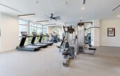 Located at 27077 S Dixie Hwy in Homestead, FL, this single-floor fitness center offers a generously sized, well-appointed exercise facility designed to support a comprehensive range of workout routines. The space is characterized by bright, white walls, light-toned carpet flooring, and modern linear pendant lighting that creates a clean, energizing atmosphere throughout.

The main strength training area is equipped with a robust selection of cable machines, selectorized strength equipment, and a dedicated free weights section featuring a full dumbbell rack lined against large wall mirrors. A power rack with adjustable benches provides space for compound lifts, while motivational wall typography — including bold "DRIVE" and "FOCUS" graphics — reinforces an active environment.

A separate cardio zone lines the perimeter, where floor-to-ceiling windows flood the space with natural light and offer open views of the surrounding landscape. Treadmills with integrated screens and additional cardio machines are positioned to take full advantage of these views.

An adjacent cycling studio accommodates indoor bikes beneath suspended linear lighting, with mirrored walls and large windows creating an open, airy feel. A functional training area features TRX suspension straps and open floor space for bodyweight and mobility work. Oversized ceiling fans provide additional ventilation throughout the facility, completing this thoughtfully designed, full-service fitness space.