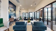 This well-appointed single-floor space is designed as a modern communal lounge and coworking area, offering a thoughtfully curated blend of collaborative and private spaces. The main lounge area features soaring ceilings and light wood-look flooring, creating an open, airy atmosphere. Bold deep-blue accent walls punctuate the cream-toned surroundings, establishing a cohesive coastal-inspired color palette throughout. Built-in shelving with an arched, botanical-wallpapered niche serves as a striking focal point, complemented by curated décor, globe sculptures, and lush greenery.

Seating is arranged to encourage both casual conversation and focused work, with richly textured blue lounge chairs, a green velvet sofa, and neutral accent seating positioned around coffee tables. A built-in workstation with multiple desk spaces and task chairs provides a dedicated area for productivity.

Glass-panel partition walls with black steel frames separate the lounge from private conference rooms. These meeting rooms feature large windows with floor-length drapes, abundant natural light, and exterior views. One room includes a round table with velvet seating beneath a dramatic fringe chandelier, while another offers a long rectangular table, mounted TV, and bold blue walls, making both suitable for formal meetings or collaborative gatherings.