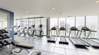 This high-rise fitness center offers a comprehensive workout environment designed to accommodate a wide range of exercise needs. The space is organized into distinct functional zones, all unified by a clean, modern aesthetic featuring a combination of white ceilings, bold blue accent walls, and durable rubber and tile flooring throughout.

The cardio zone is positioned along a wall of floor-to-ceiling windows, providing sweeping views of the surrounding city skyline. Multiple treadmills, elliptical trainers, and cycling machines are arranged to allow users to exercise while taking in the urban panorama, with ample natural light filling the area.

Adjacent to the cardio section, the strength training zone features a robust selection of equipment, including free weight benches, barbells, weight plates, cable machines, and a multi-functional Smith machine. Bold blue walls and large wall-mounted mirrors define this area, creating an energizing atmosphere. Ceiling fans provide added airflow and comfort.

A dedicated functional training area rounds out the facility, equipped with kettlebells in a range of sizes, medicine balls, resistance tools, and open floor space for stretching and bodyweight exercises. Large sliding glass doors in this zone open to views of outdoor greenery, enhancing the sense of openness. The overall layout is spacious, well-lit, and thoughtfully arranged to support a complete fitness routine.