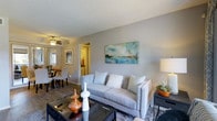 This single-floor residence is designed with an open-concept layout that seamlessly connects the main living areas. The living room serves as the central gathering space, featuring wood-style flooring, neutral gray walls, and a textured ceiling. A glass-top coffee table and a plush gray sofa anchor the room, while a framed abstract canvas in blue and teal tones adds a decorative focal point to the wall. A ceiling fan with integrated lighting provides both comfort and illumination.

The open flow continues into the dining area, which is positioned adjacent to the living room and benefits from natural light streaming through a nearby window. Dining chairs surround a modest table, creating an inviting space for meals. Beyond the dining area, a hallway leads to the kitchen, which features yellow-toned cabinetry and additional lighting. The consistent wood-style flooring throughout the main living areas ties the spaces together, contributing to a cohesive and functional layout. The overall ambiance is modern, clean, and comfortable, making efficient use of the available space.