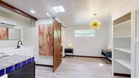 Located at 239 N Warren Ave in Tucson, Arizona, this efficiently designed single-floor studio makes a strong architectural statement despite its compact footprint. The open-plan living area combines dining, kitchen, and living functions in a cohesive layout anchored by light wood-look flooring and crisp white walls throughout.

A standout feature is the full-height rustic sliding barn door crafted from rich, warm-toned wood, which adds a bold rustic-modern contrast to the otherwise contemporary interior. Above, a dramatic gold sputnik chandelier serves as a focal centerpiece, while a skylight draws in additional natural light from above. Exposed dark wood ceiling beams further reinforce the space's character.

The kitchenette is outfitted with white cabinetry, a wall-mounted mini-split climate control unit, and a distinctively decorative Talavera tile countertop in blue and white, adding a nod to regional Southwestern style. Built-in open shelving alongside a louvered closet door provides practical storage within the living area.

The bathroom is clean and well-appointed, featuring a walk-in shower clad in classic white subway tile with dark grout accents, a sleek dark-framed glass enclosure, and a window that invites natural light. The overall ambiance of the space is modern yet warm, blending rustic and contemporary design elements with functional, space-conscious planning.