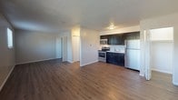 This single-floor space offers a clean, functional layout designed for comfortable everyday living. The open-concept main area seamlessly connects the living space to the kitchen, creating an airy and versatile environment. The kitchen is well-appointed with dark navy cabinetry, a stainless steel refrigerator, electric range, and over-the-range microwave, all set against light-toned countertops and a single-basin sink. Rich, warm-toned wood-look flooring runs consistently throughout the space, adding a sense of cohesion and warmth. Textured ceilings and crisp white trim complement the neutral wall tones throughout. A hallway off the main living area leads to the remaining rooms, including a generously sized bedroom featuring two windows with horizontal blinds that invite natural light and outdoor views, creating a bright and welcoming retreat. The bathroom includes a full-size soaking tub with an integrated shower surround in white, paired with brushed nickel fixtures for a clean, modern finish. Closet space is accessible from the hallway, providing practical storage. Overall, the space presents a straightforward and well-maintained interior suited for a range of occupants.