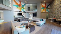Located at 10501 Landing Way in Miamisburg, Ohio, this dynamic single-floor community lounge is designed to foster connection and comfort through bold aesthetics and thoughtfully arranged spaces. The centerpiece of the room is a double-height ceiling that amplifies the sense of openness, adorned with large-scale, colorful murals depicting outstretched hands — a striking artistic statement that sets an energetic and welcoming tone throughout. The main lounge area features generous sectional seating in light gray tones, accented with teal and multicolor throw pillows, arranged around low coffee tables on a neutral area rug. Wood-look plank flooring with inlaid geometric carpet accents runs throughout, grounding the space with warmth. A dedicated kitchenette area provides a sink, dark cabinetry with stainless steel appliances, a coffee machine, and bar-height seating — ideal for casual refreshments. A secondary lounge zone offers curved tulip-style chairs and round marble-top tables set against a vivid, multi-colored abstract wallcovering, creating an intimate nook for conversation or individual use. Recessed lighting and wall-mounted flat-screen televisions enhance functionality, while floor-to-ceiling glass entry doors invite natural light from the exterior. The overall ambiance is modern, lively, and community-oriented.