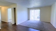 This single-floor residential space offers a clean and functional layout designed for comfortable everyday living. The main living area is a standout feature, bathed in natural light that streams through large sliding glass doors fitted with vertical blinds, opening to an outdoor area and providing pleasant views of the surrounding greenery. The room showcases a combination of dark hardwood-style flooring at the entry transition and soft, plush carpeting throughout the main living space, creating a warm and inviting atmosphere. Neutral, light-toned walls complement the overall brightness of the interior, while white baseboards add a crisp finishing touch. The ceiling features a classic textured finish, adding subtle depth to the space. A paneled interior door visible to the side hints at additional rooms beyond, suggesting a practical and well-organized floor plan. The overall ambiance is airy and understated, with an emphasis on natural light and simplicity, making it a versatile space well-suited for a variety of furnishing styles and living arrangements.