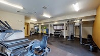 This fitness center offers a comprehensive workout environment designed to accommodate a variety of exercise routines. The space is finished with durable dark rubber flooring, providing both safety and comfort during workouts. Overhead fluorescent lighting, supplemented by recessed fixtures, ensures the area is well-lit throughout.

Cardio equipment, including a treadmill and a recumbent bike, is positioned toward the front of the room, offering accessible options for warm-ups or cardiovascular training. A multi-station cable machine anchors one side of the room, enabling a wide range of strength-training exercises. A well-stocked dumbbell rack holds an organized selection of free weights, catering to users of varying fitness levels.

Large mirrors line one wall, enhancing the sense of space while allowing users to monitor their form. Exercise and stability balls add versatility for functional training. A window with a blind allows for natural light, contributing to an open, energizing atmosphere. A secondary area visible through a doorway suggests additional utility space, such as changing facilities or storage.