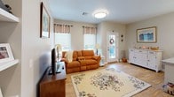 This single-floor residence offers a thoughtfully designed open layout with warm neutral tones and hardwood flooring throughout the main living areas. The kitchen serves as a functional centerpiece, showcasing granite countertops, white cabinetry, stainless steel appliances including a refrigerator, microwave, and range, and a peninsula with pendant lighting overhead. A window above the sink allows natural light to filter in, while open shelving provides additional storage. Adjacent to the kitchen, a dedicated dining area accommodates a wood table and chairs beneath a large decorative photo collage wall display. The living room features hardwood flooring, a comfortable seating arrangement, and windows dressed with valances and roller shades that allow for adjustable natural light. A flexible secondary room functions as a home office and sitting area, benefiting from dual windows and carpeted flooring. The bedroom is well-proportioned with carpeted floors, a ceiling fan, and two windows with curtains. The primary bathroom is finished with a granite vanity, white cabinetry, and a glass-enclosed walk-in shower with tile surround. A second, more compact bathroom provides a toilet and tile flooring.