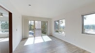 This single-floor space offers a clean, contemporary design characterized by an open and airy atmosphere. The main living area is bathed in natural light, courtesy of large sliding glass doors that open onto a private balcony overlooking lush, mature trees. A generously sized picture window on the adjacent wall further amplifies the sense of openness while framing tranquil wooded views from multiple angles.

The interior features smooth white walls and ceilings that reflect natural light throughout the day, enhancing the bright and welcoming ambiance. Light-toned hardwood-style flooring runs throughout the living area, contributing warmth and a sense of continuity to the space. The sliding door leads to a slatted balcony railing, providing both privacy and an inviting outdoor extension of the living area.

The layout is thoughtfully designed to maximize both comfort and functionality, with a spacious feel that accommodates a variety of furniture arrangements. The minimalist aesthetic, combined with an abundance of natural light and verdant outdoor views, creates a calm and pleasant environment. The space presents a well-maintained, move-in-ready condition with modern finishes that appeal to a wide range of tastes.