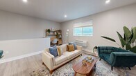 This two-floor unit offers a thoughtfully designed layout that maximizes functionality and comfort. The main living level, which comprises the majority of the floor area, is accessed via a carpeted staircase with a sleek white handrail, leading up to a bright and open living space. The combined kitchen and living room forms the heart of the unit, featuring light hardwood-style flooring that runs seamlessly throughout. The kitchen is well-equipped with white shaker-style cabinetry, granite countertops, stainless steel appliances including a refrigerator, range, and microwave, and a kitchen island with a deep sink. The living area is open and airy, with recessed lighting, a large window that allows ample natural light and outdoor views, and enough space to accommodate a comfortable seating arrangement alongside a dedicated workspace. The bathroom is finished with light hardwood-style flooring, white cabinetry with brushed hardware, a granite-topped vanity, a large mirror, and clean neutral walls that create a calm, spa-like atmosphere. The overall design palette is modern and cohesive, combining neutral tones, contemporary fixtures, and quality materials to deliver a polished and functional living environment.