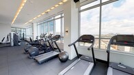 This contemporary fitness center is designed to deliver a premium workout experience with an abundance of natural light and expansive city views. Floor-to-ceiling glass windows wrap around the space, offering sweeping panoramic vistas of the surrounding urban skyline that serve as an energizing backdrop during exercise. The facility is organized into distinct functional zones to accommodate a wide range of fitness activities. The cardio area features a row of treadmills and elliptical machines positioned directly in front of the large windows, allowing users to enjoy the scenery while training. A dedicated strength and resistance zone houses a comprehensive selection of cable machines, weight benches, and selectorized equipment, complemented by warm wood accent walls and stylish pendant lighting that add a refined aesthetic contrast to the athletic setting. Additional areas provide space for flexibility training and functional movement, with step platforms and medicine balls available for varied workout routines. Dumbbell racks and free weights offer further options for strength training. The flooring throughout is durable rubber tile, practical for high-impact use. Recessed and cove lighting creates a bright, motivating atmosphere. An adjacent outdoor terrace, visible through the glass walls, extends the experience beyond the interior. The overall design balances functionality with a sophisticated, modern aesthetic.