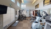 This fitness center is housed in a single-level space distinguished by dramatically high ceilings and an airy, open atmosphere. Clerestory windows positioned near the roofline, combined with large standard windows fitted with blinds, flood the interior with natural light and provide views of the surrounding outdoor area, including what appears to be a pool. The flooring throughout consists of durable, neutral-toned carpet tiles well-suited for a high-traffic exercise environment.

The cardio zone features multiple treadmills and elliptical machines by Landice, complemented by a wall-mounted flat-screen television for entertainment during workouts. A dedicated stretching and flexibility area includes yoga mats and interlocking foam floor tiles. Functional training accessories such as a stability ball, medicine ball rack with assorted weighted balls, and resistance bands are also available.

The strength training section is equipped with a multi-station cable machine, an adjustable weight bench, and a full dumbbell rack with varying weights, all positioned in front of a large wall mirror that enhances the sense of space. A posted fitness center rules board, a wall clock, and a vending machine near the entrance add practical convenience. Ceiling-mounted lighting fixtures and a portable fan ensure a comfortable workout environment year-round.